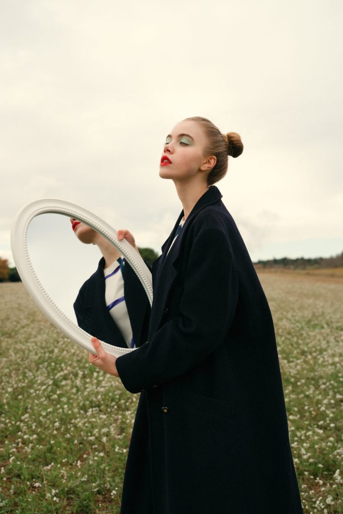 Editorial de moda: mujer con abrigo negro sosteniendo un espejo en un campo