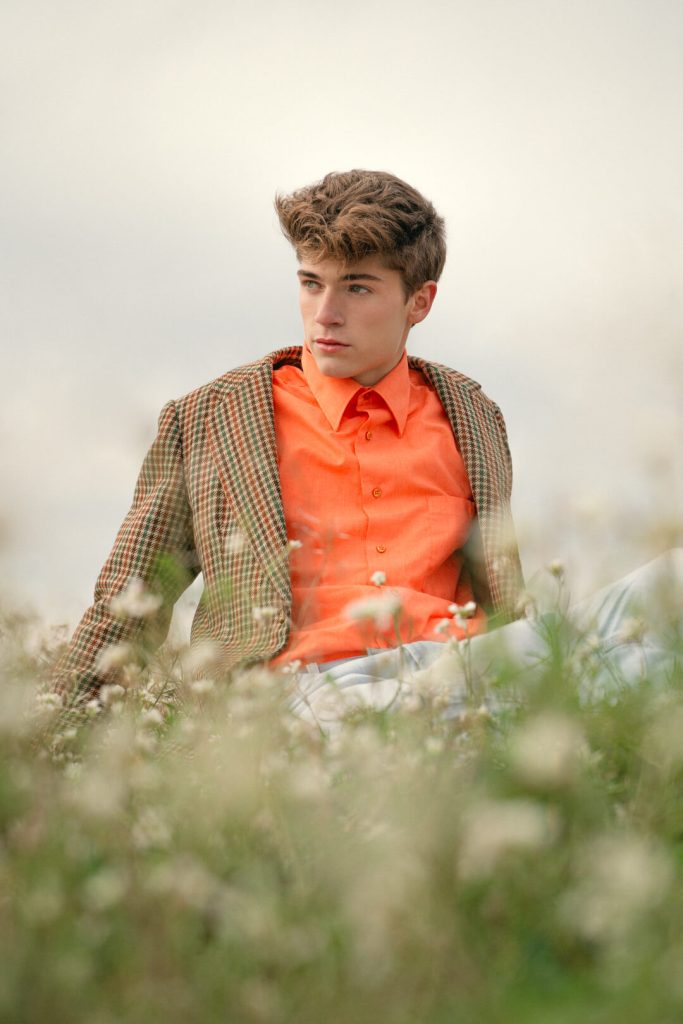 Chico con camisa naranja y chaqueta posando en un campo de flores