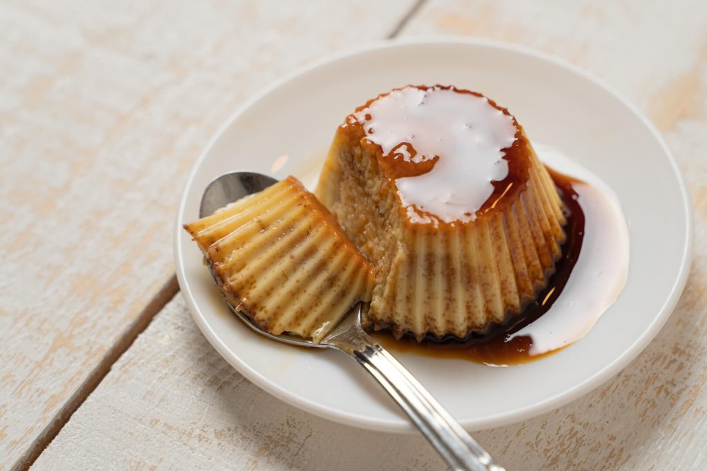 Flan con una cuchara y caramelo en un plato blanco sobre mesa de madera clara