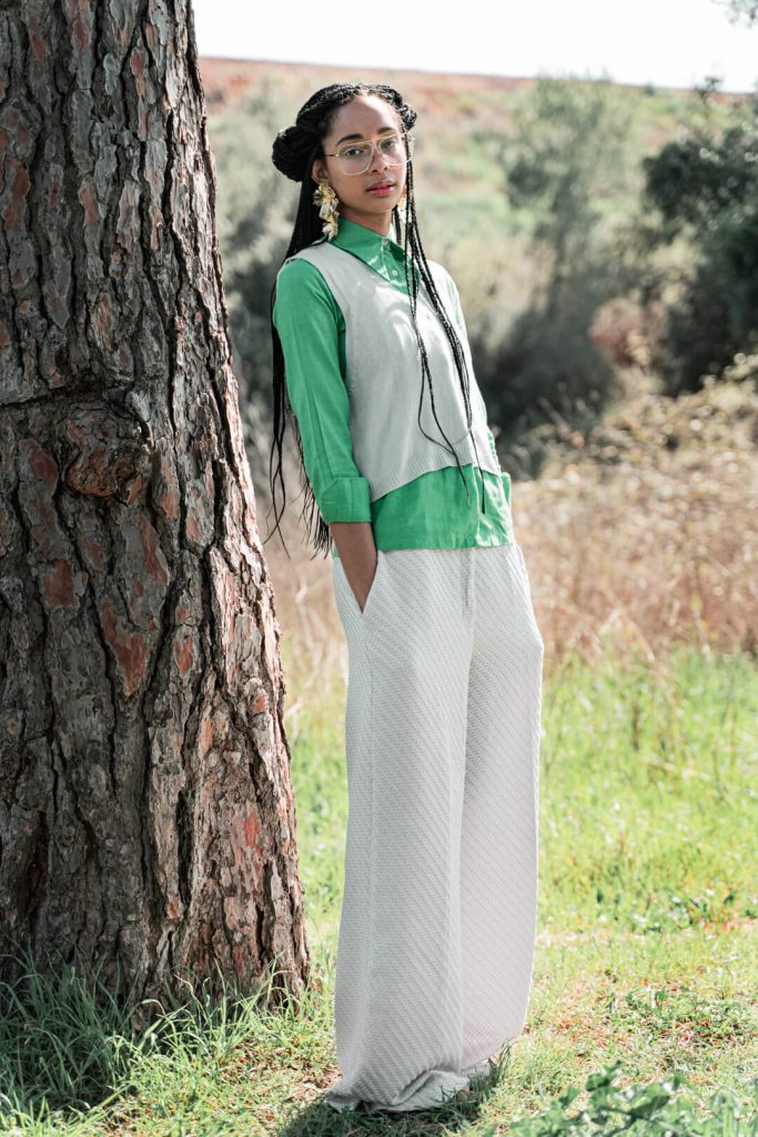 Plano de cuerpo entero de una mujer con chaleco verde y pantalón blanco junto a un árbol