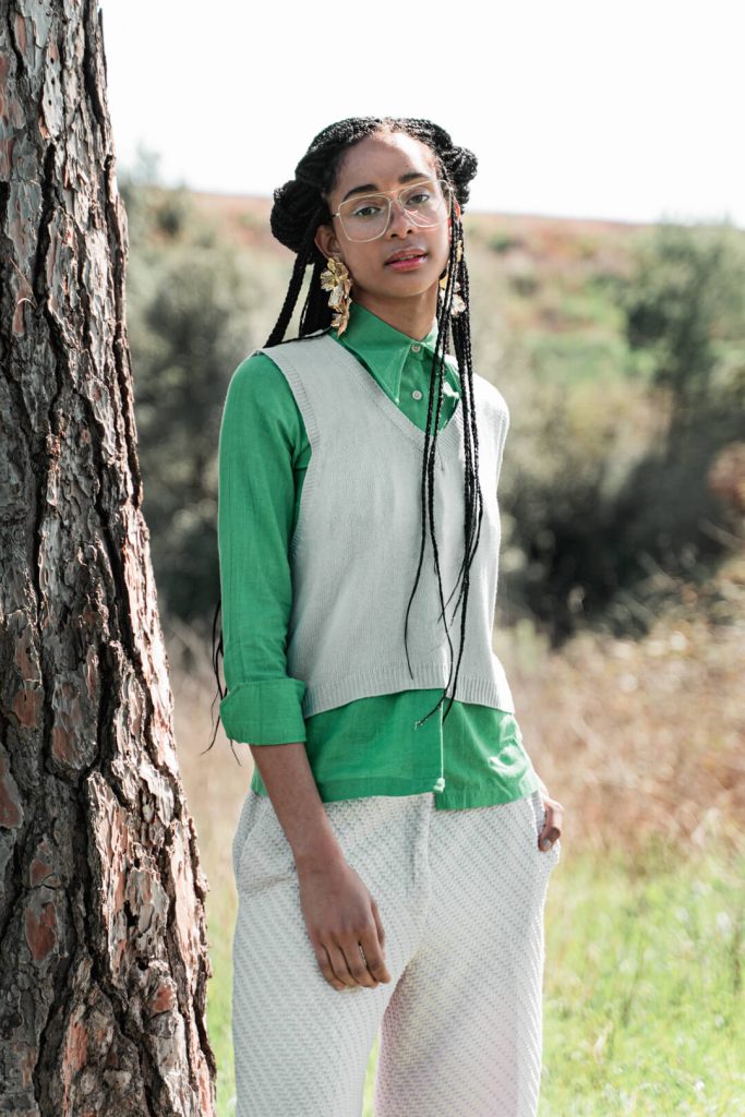 Mujer con chaleco verde y pantalón blanco posando junto a un árbol