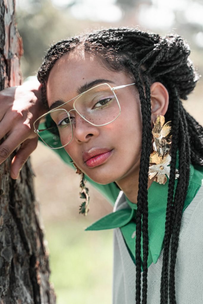 Retrato de moda de una mujer con gafas, trenzas y pendientes junto a un árbol