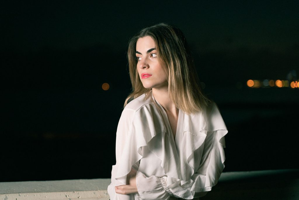 Retrato nocturno de una mujer con blusa blanca apoyada en una barandilla con luces de fondo