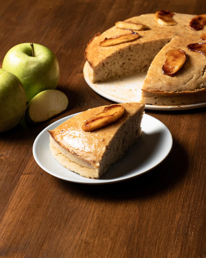 Porción de tarta de manzana en un plato con la tarta entera detrás, fotografía gastronómica vertical